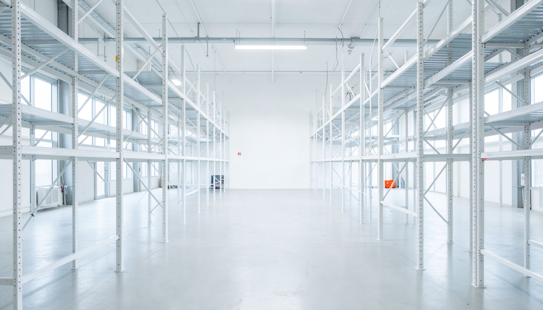 Clean symmetrical architectural photograph of a bright distribution warehouse interior with white steel shelving, pale concrete floor, and soft natural light from tall windows, in cool white and steel grey tones.