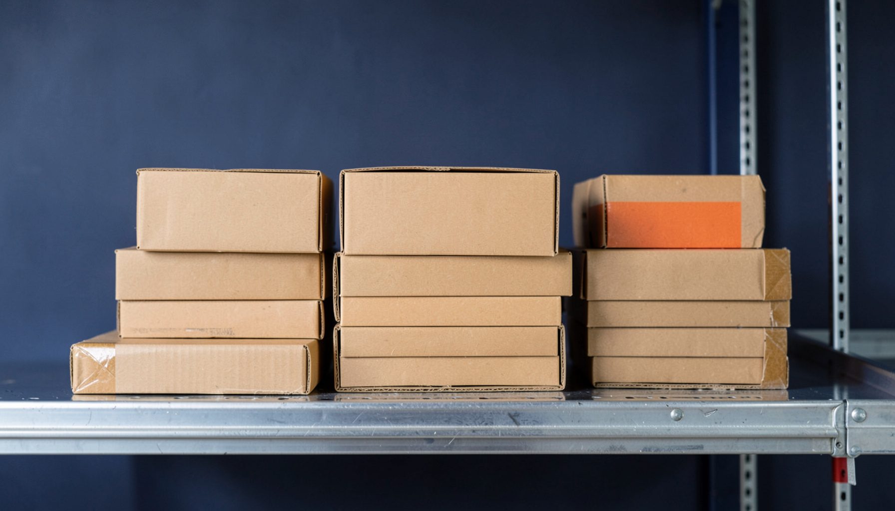 Stacked cardboard parcels ready for dispatch on a metal warehouse shelf, lit with neutral daylight and a soft navy backdrop.