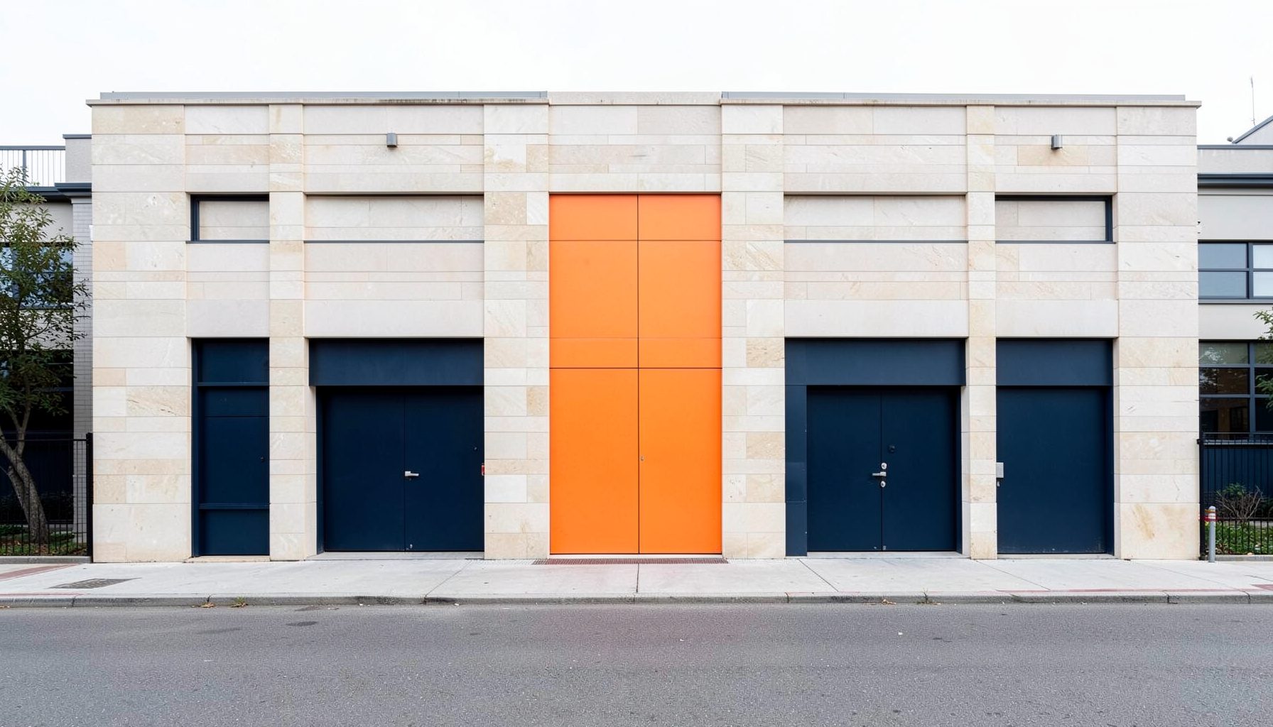Symmetrical architectural exterior of a modern light-industrial unit in pale stone with dark navy doors, set behind a quiet street.