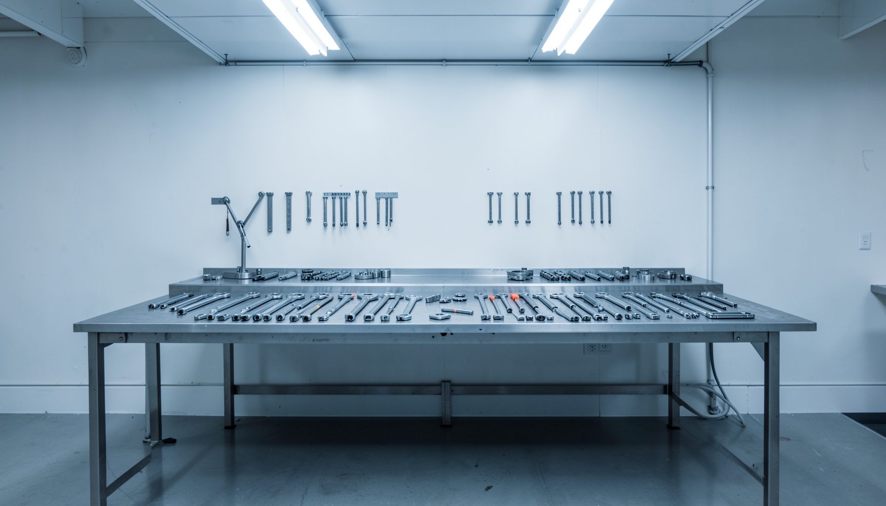 Symmetrical photograph of a clean workshop bench with neatly arranged metal tools and parts under cool white light, conveying an orderly, technical mood with grey and steel-blue tones.
